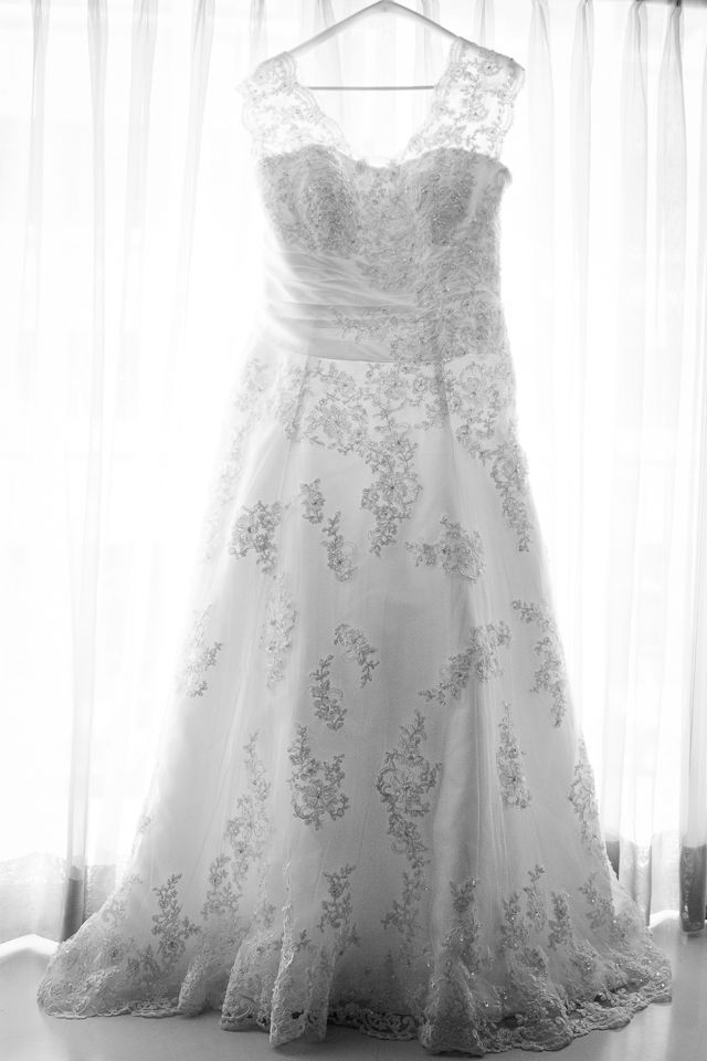 A white lace A-line wedding dress with a V-neckline hangs on a hanger in front of a window with white curtains.