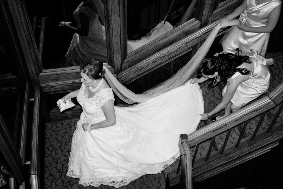 A bride in a white gown walks down a wooden staircase while two assistants hold up the long train of her dress.