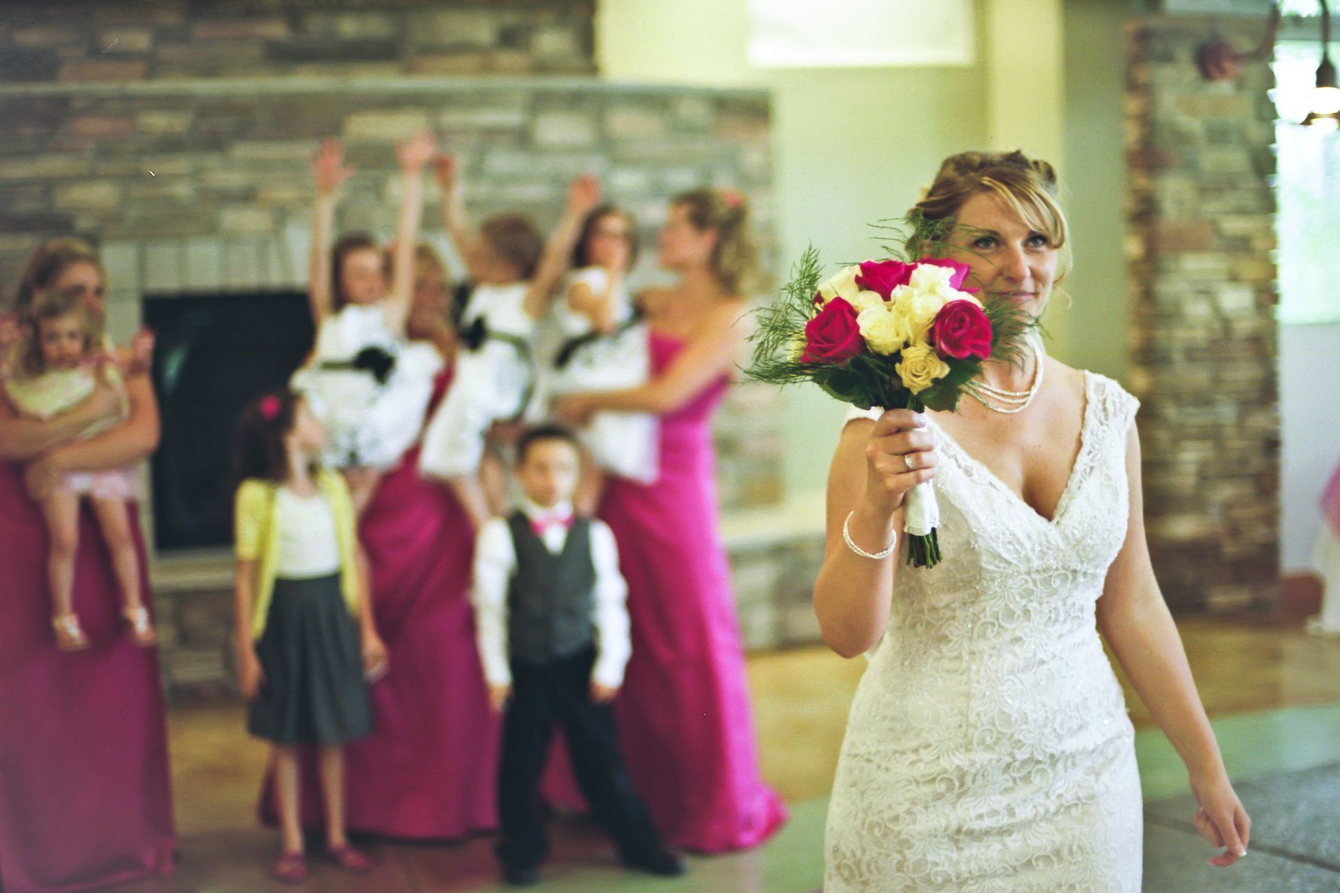 A bride in a white wedding dress holds a bouquet, with blurred wedding guests standing behind her in a rustic venue.