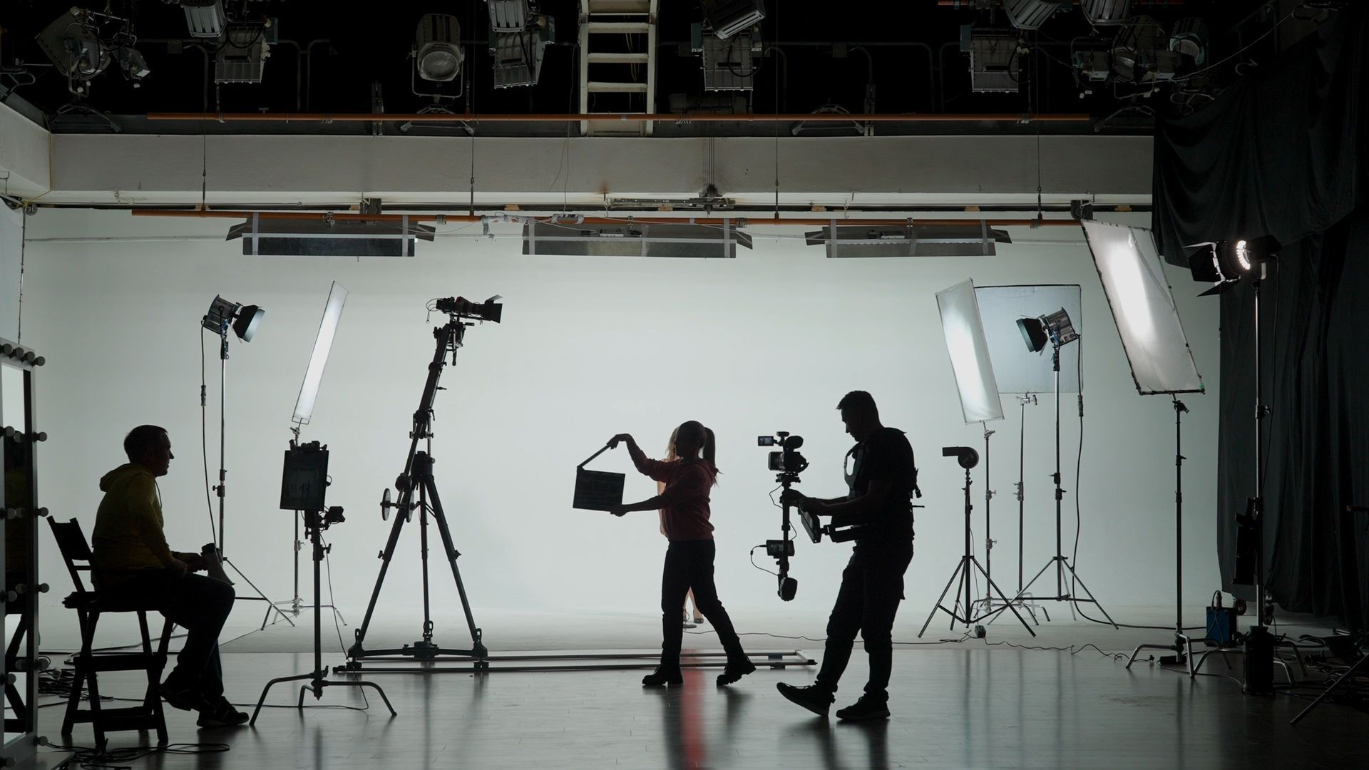 Film crew in a studio, silhouetted against a white backdrop with lights and cameras.