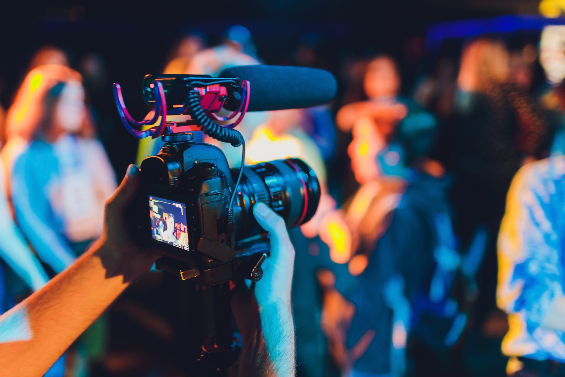 Person holding a camera with a microphone recording a blurred crowd at a venue.