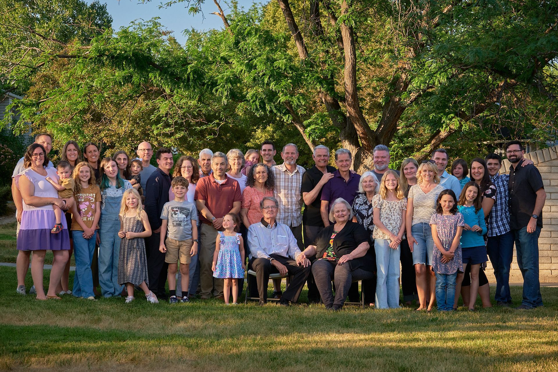 A large, multi-generational family poses for a portrait outdoors in front of a tree at golden hour.