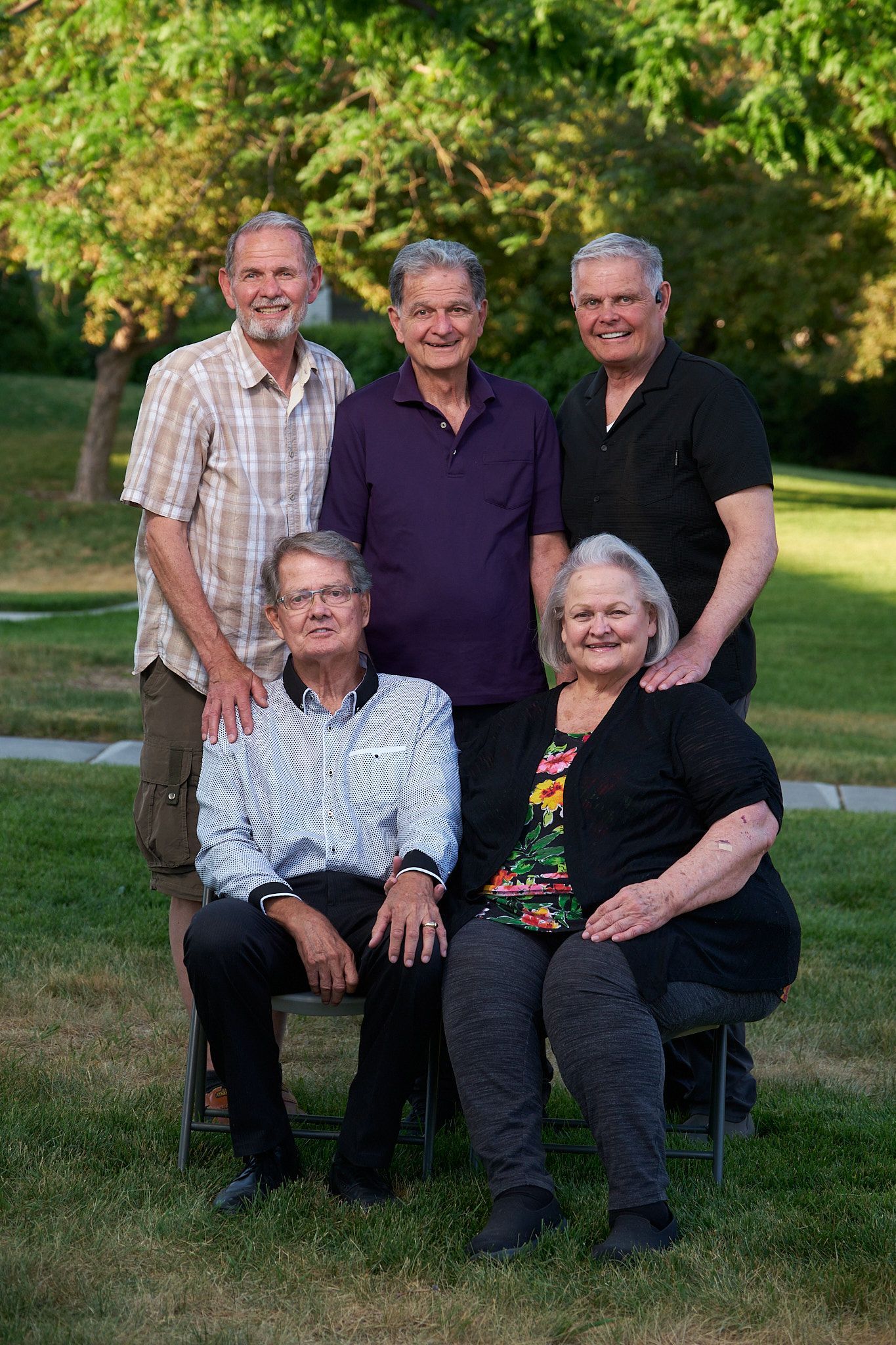 A family portrait of five people standing and sitting on chairs in a green, grassy park setting.