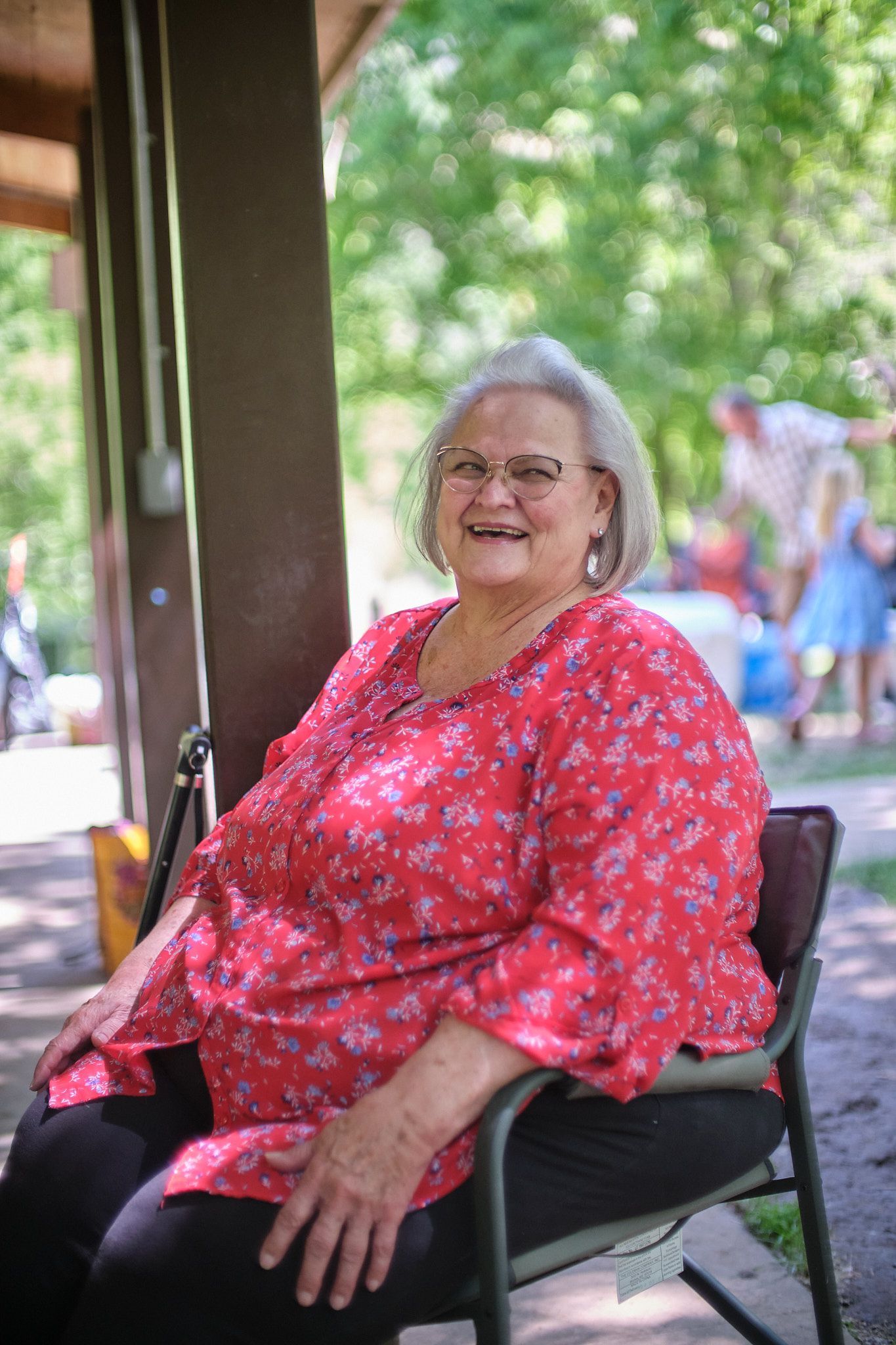 An individual with gray hair wearing a red floral blouse and black pants, sitting in a chair outdoors.