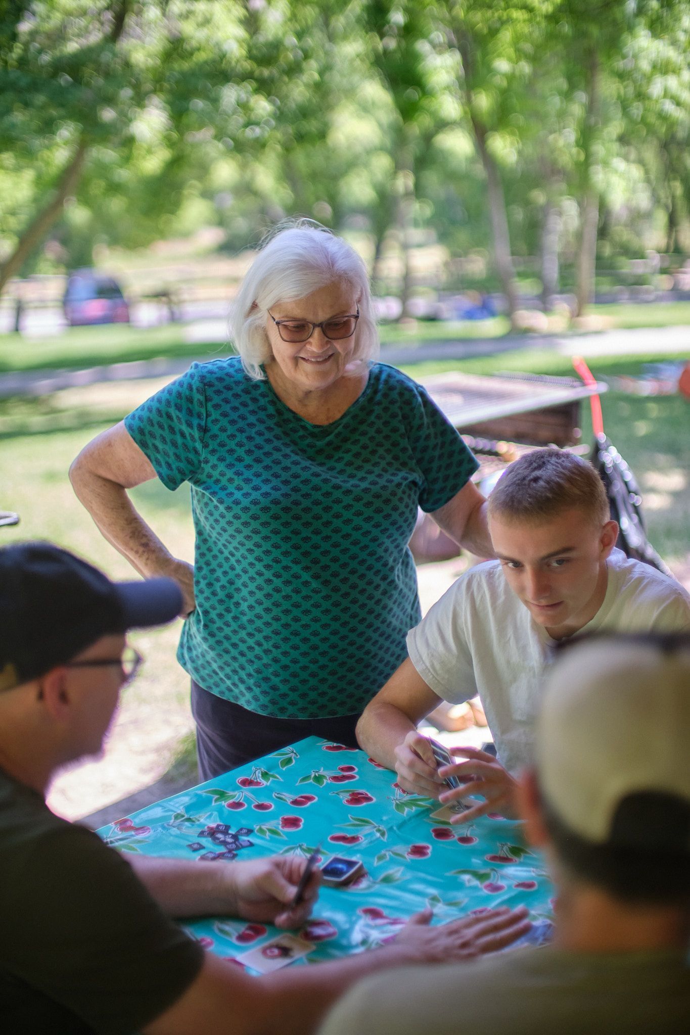 Four people sit around a table at a park, playing a card game on a patterned, teal tablecloth.