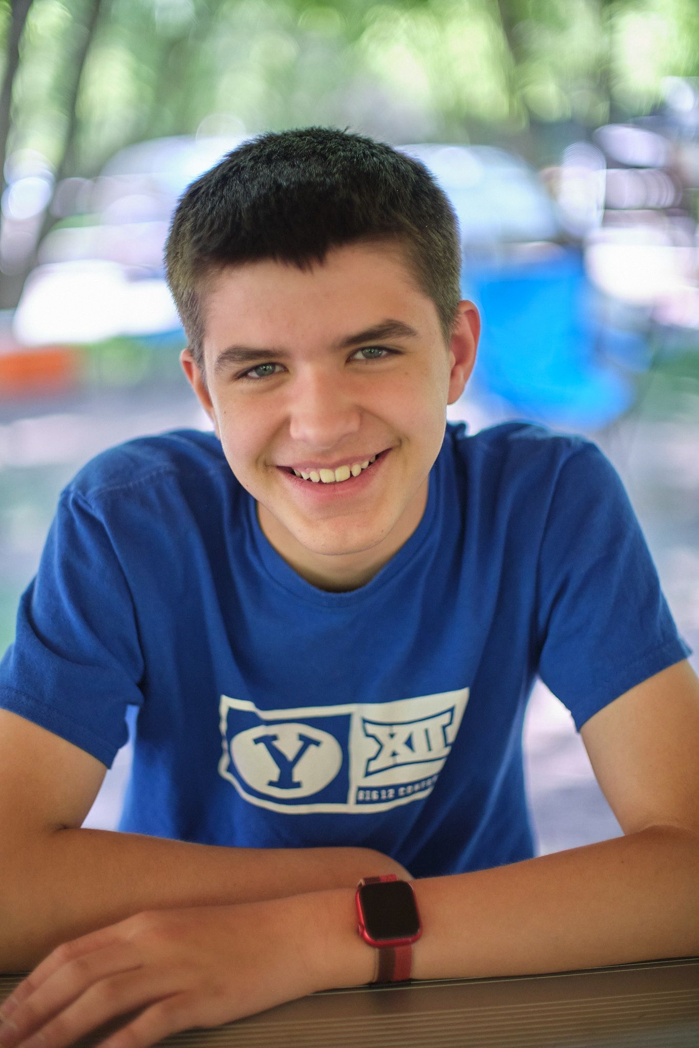 A smiling person in a blue BYU t-shirt with a smartwatch sits outdoors in a soft-focus green setting.