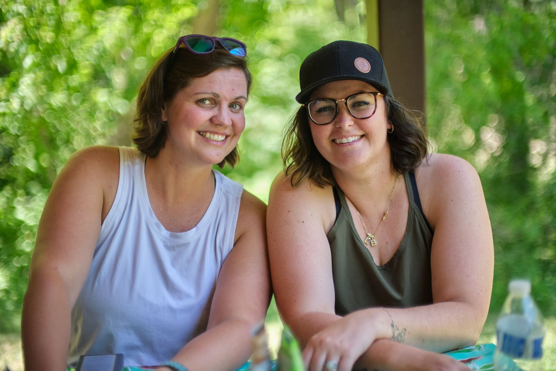 Two smiling people sitting side-by-side outdoors, wearing tank tops and glasses, set against a blurry green background.