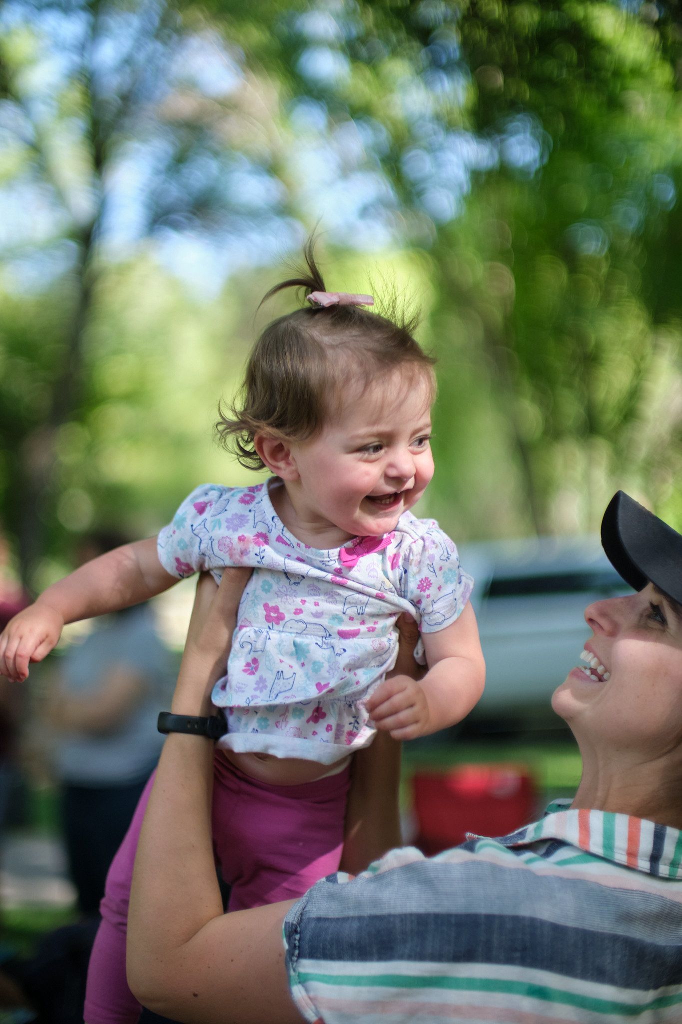 A person in a striped shirt and black hat lifts a laughing toddler in a floral top and pink pants outdoors.