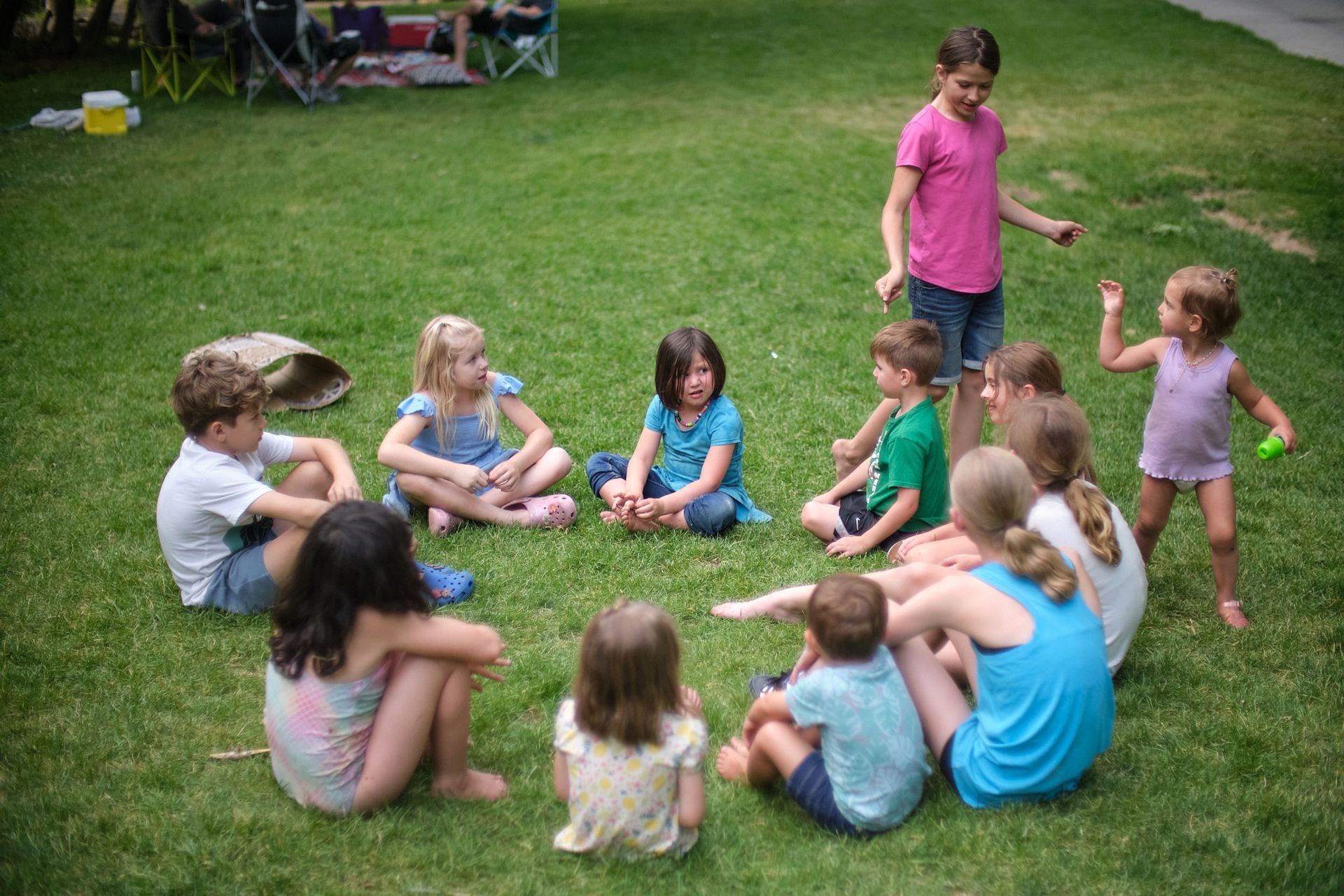 A group of children sits in a circle on green grass, with one child standing nearby in a bright pink shirt.