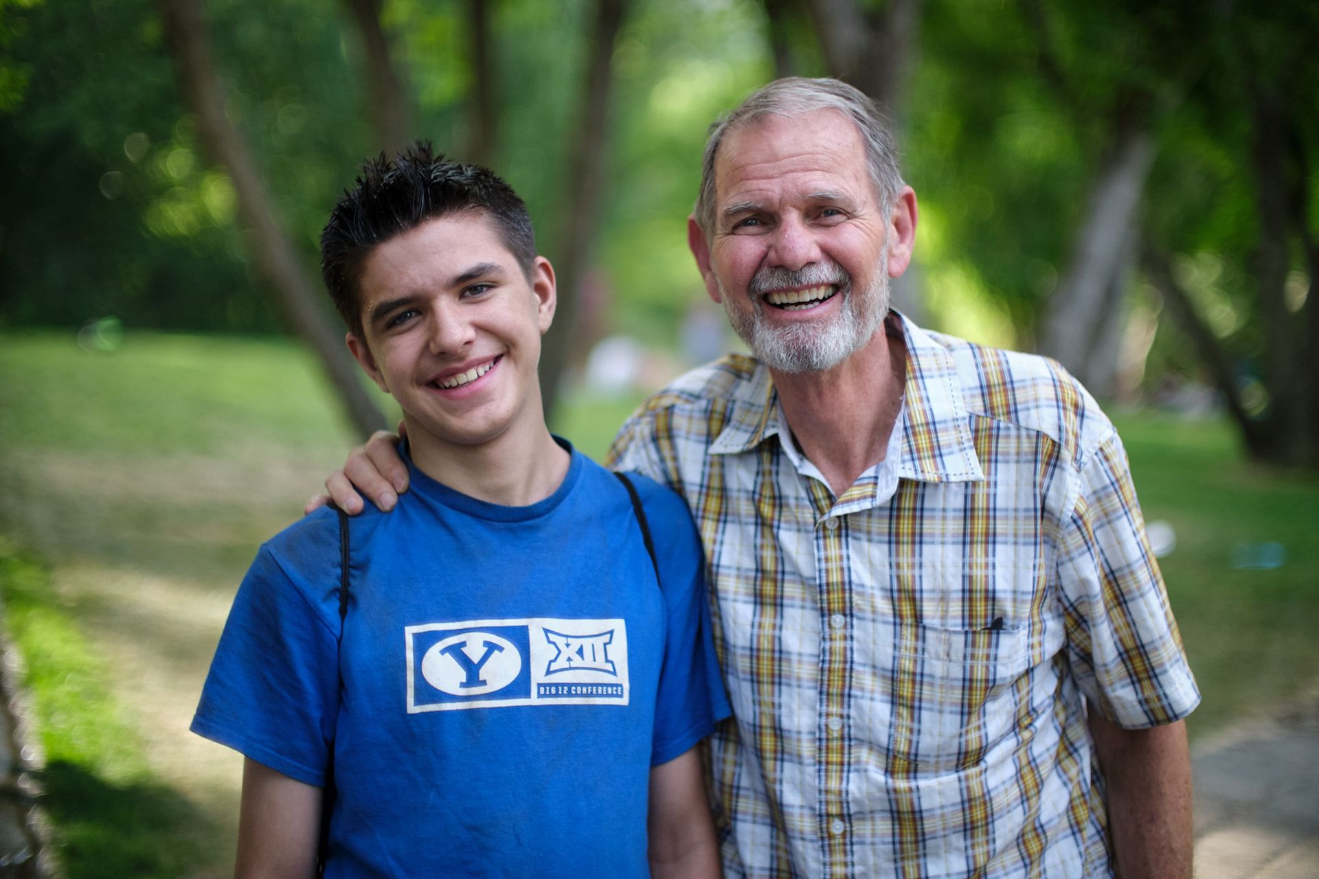 Two smiling people stand together outdoors with a blurred park background; one wears a blue BYU t-shirt, the other a plaid.