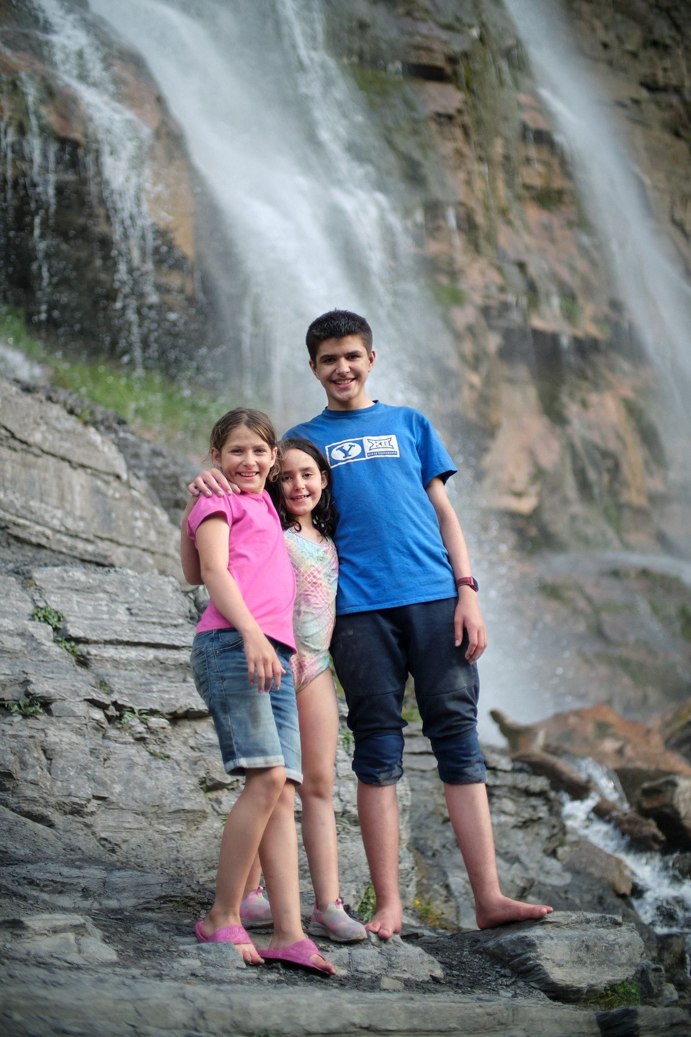 Three people standing together on a rocky path in front of a waterfall, smiling at the camera.