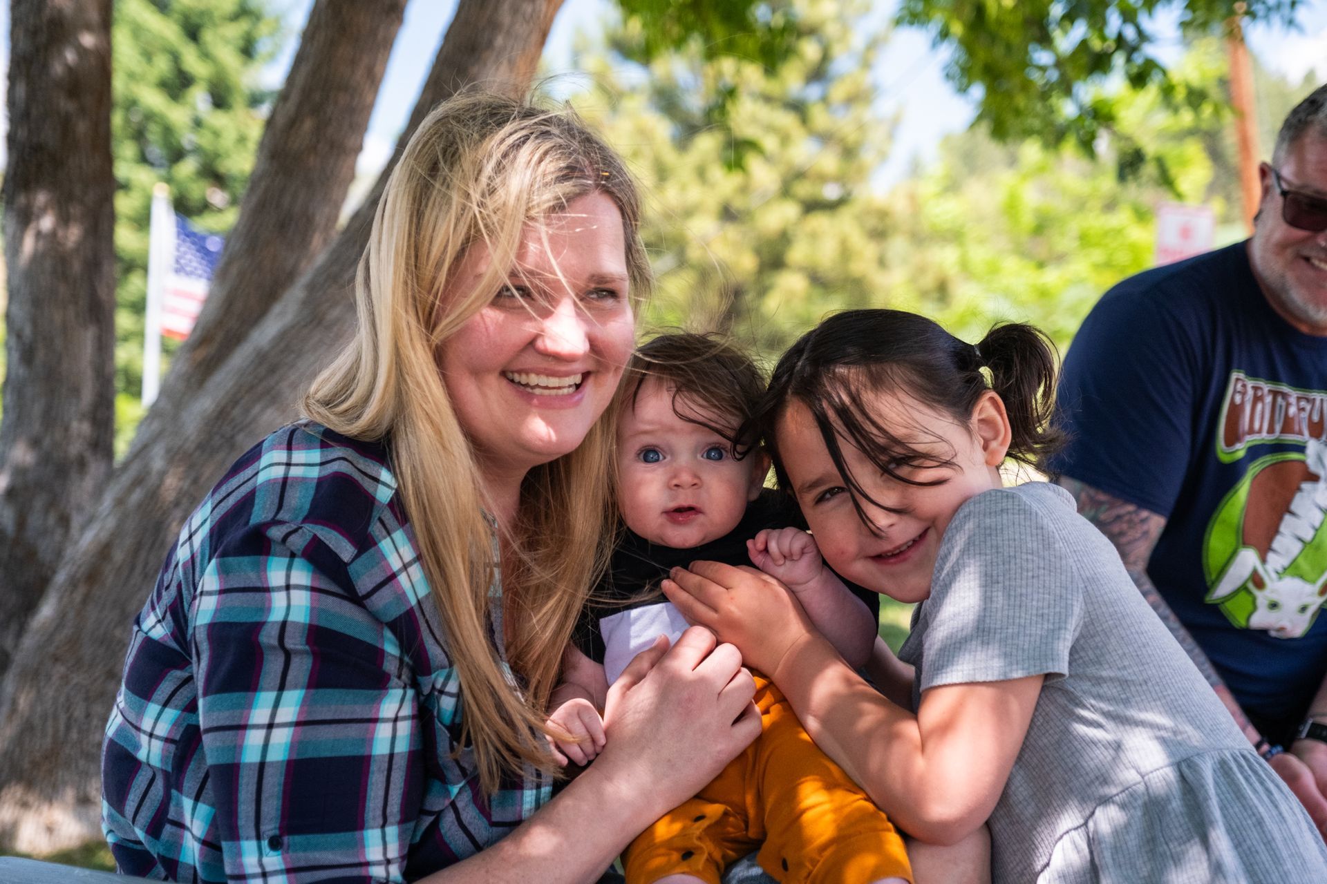 A woman and child hold an infant in an outdoor setting, with a man visible in the background.