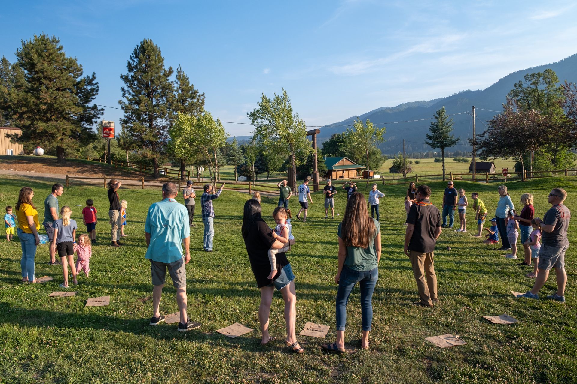 A diverse group of people stands in a large circle on a grassy field outdoors, with mountains in the background.