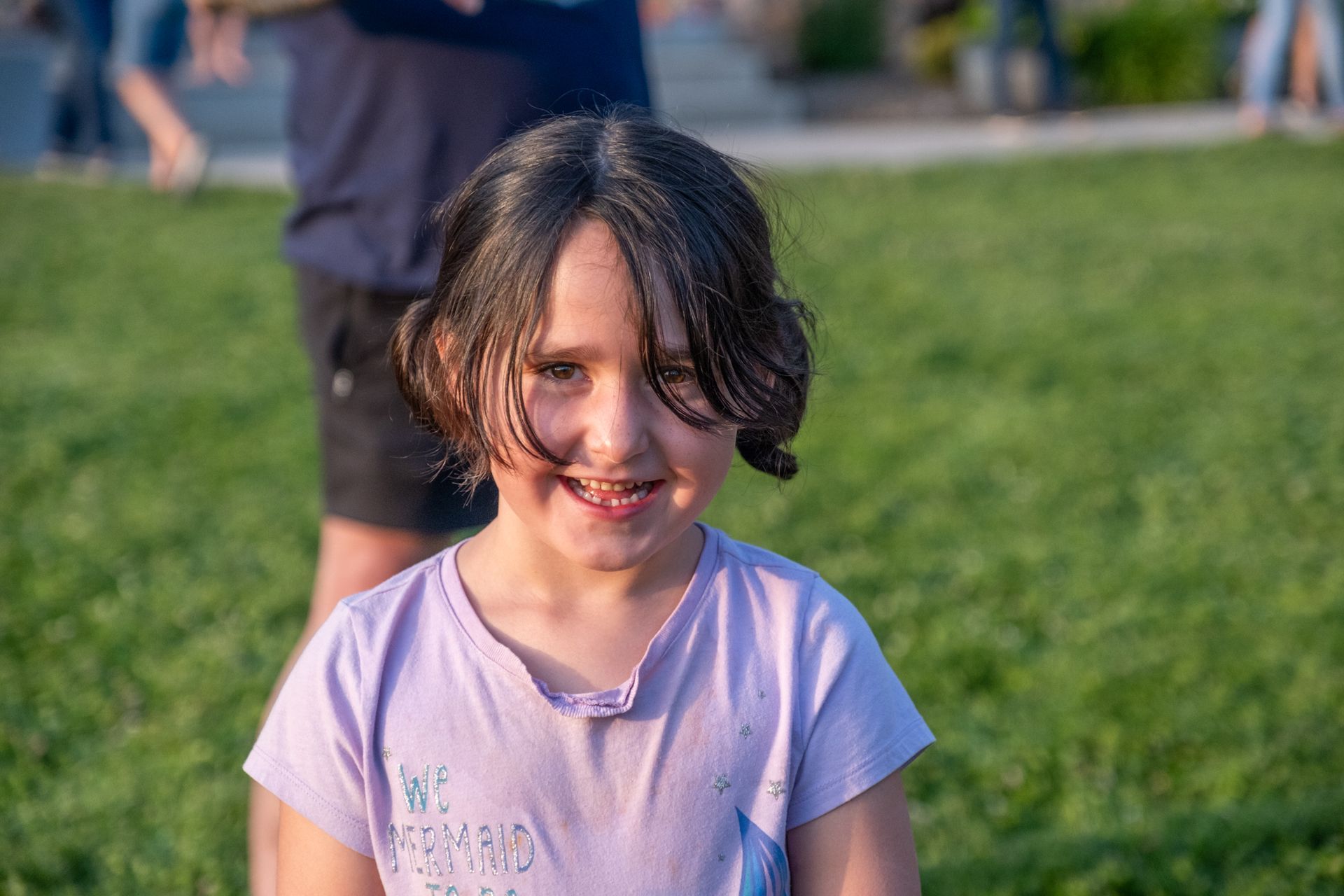 A smiling child with dark hair in a light purple t-shirt stands outside on a green lawn.