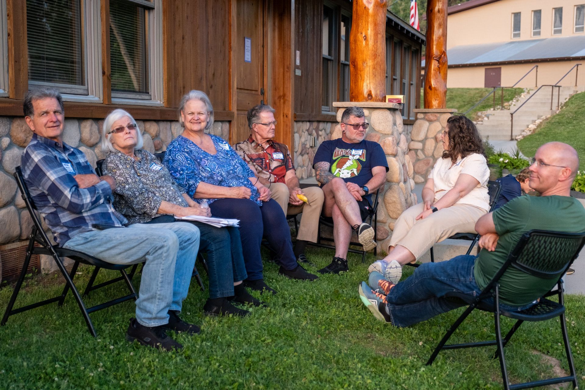 Six people sit in a circle on folding chairs outside a log cabin, talking during a casual outdoor gathering.