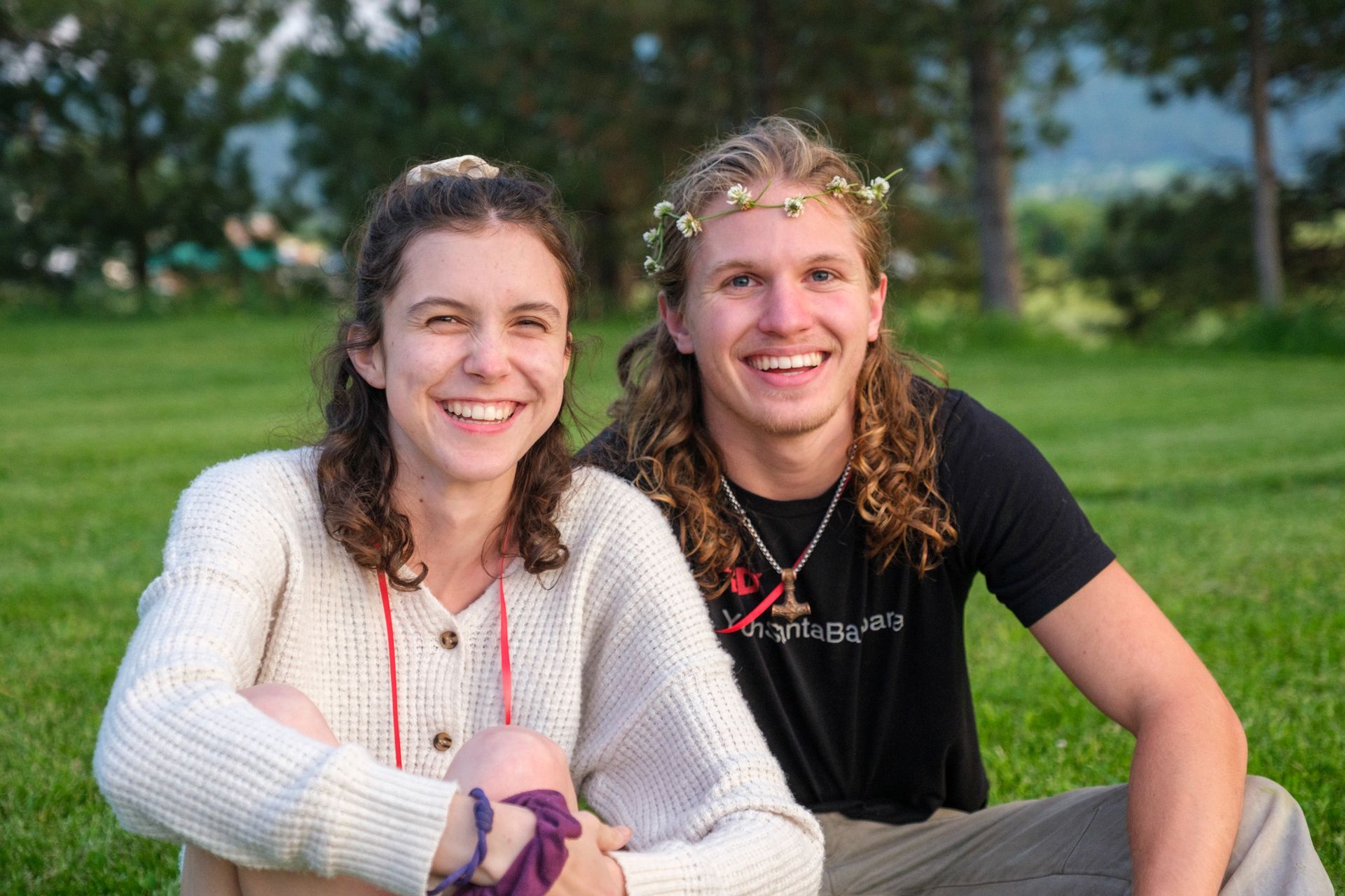 Two smiling people sitting on a grassy lawn outdoors, the person on the right wearing a small floral crown.
