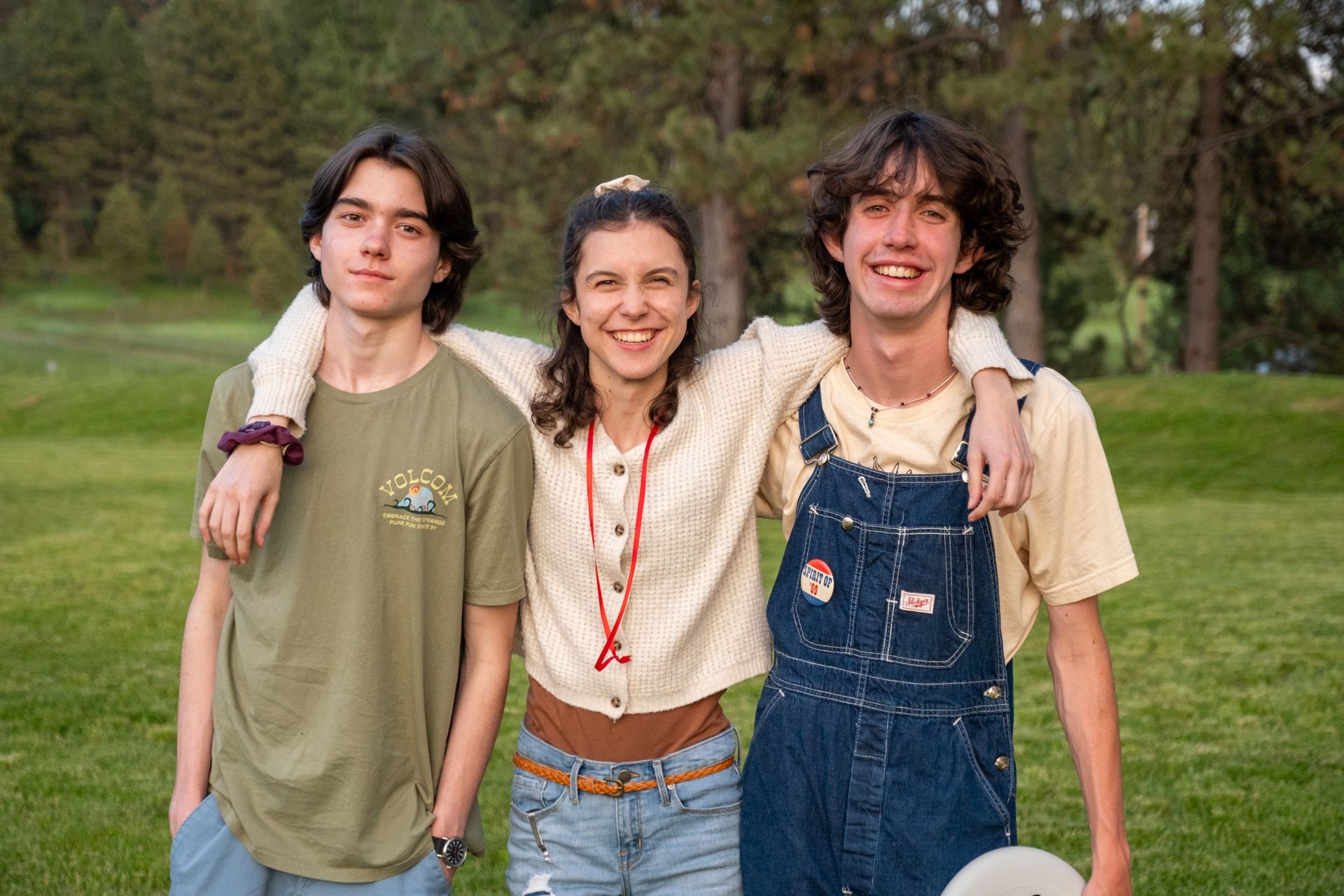Three people stand together smiling, arms around each other's shoulders, in a grassy outdoor setting with trees behind.