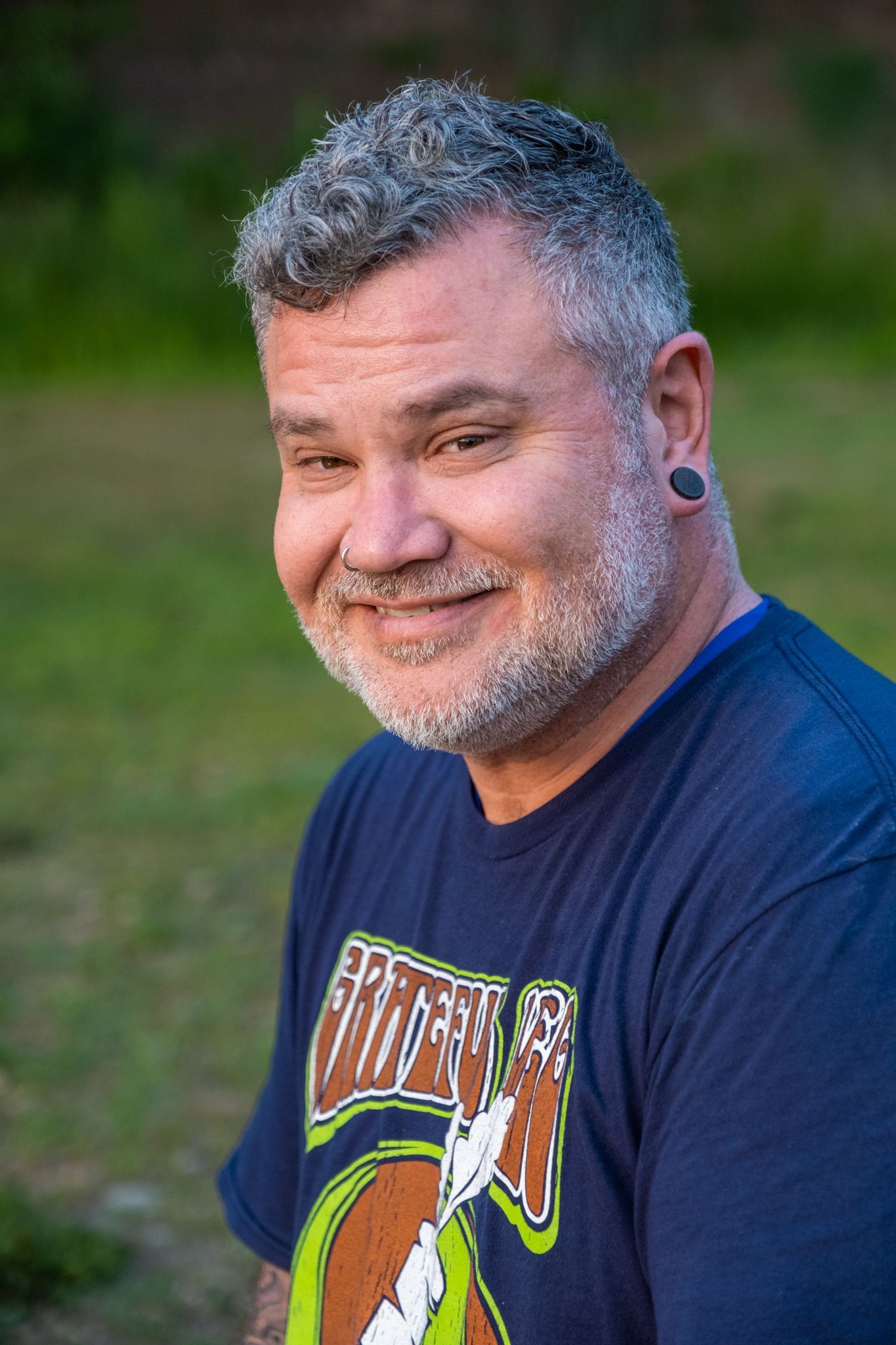 A smiling individual with salt-and-pepper hair and beard, wearing a graphic t-shirt in an outdoor, grassy setting.