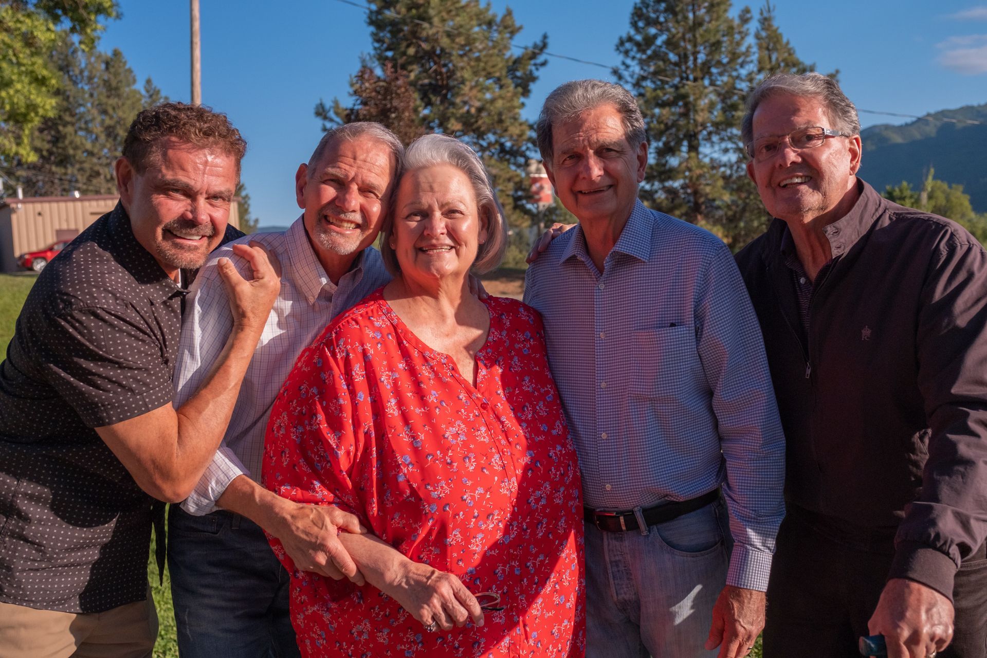 Five people smile together outdoors in a sunny park setting, standing in a line with trees and a mountain in the background.