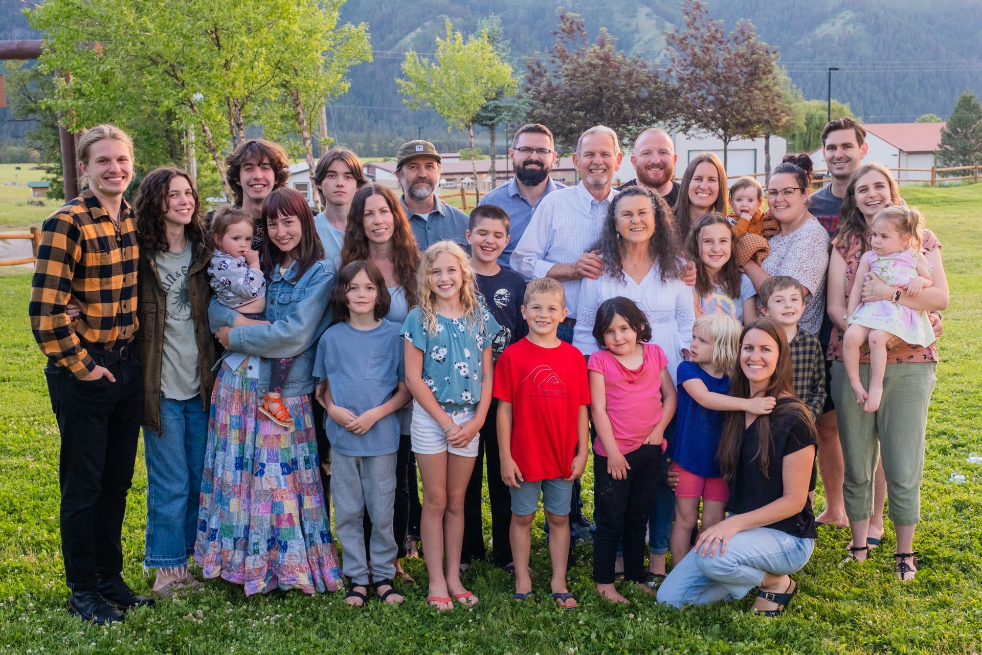 A large, smiling family poses for a photo outdoors in a grassy field with mountains in the background.