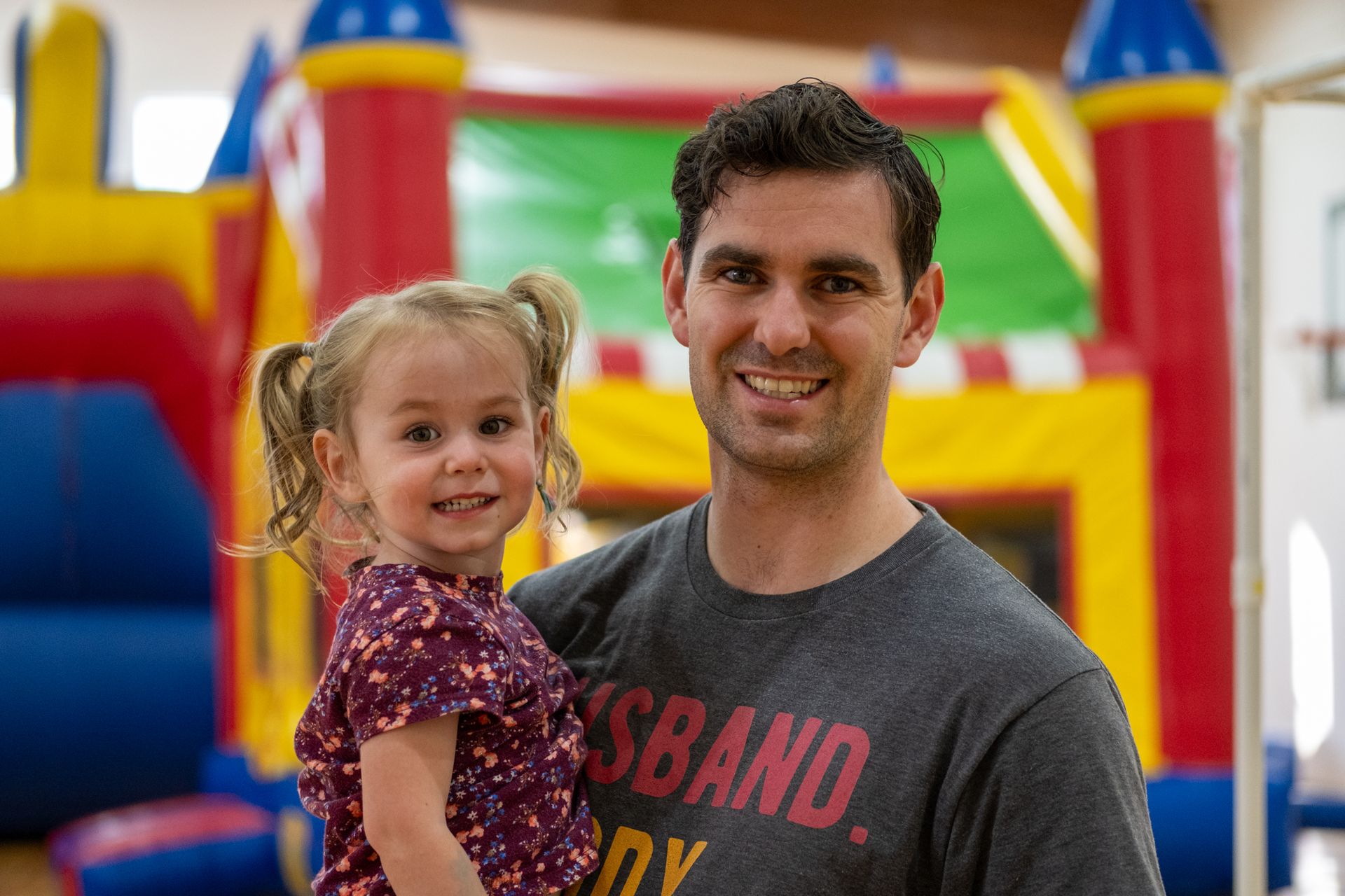 A person smiling while holding a young child in front of a colorful indoor bounce house.
