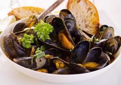Una ciotola bianca piena di cozze cotte guarnite con erbe aromatiche, servita con fette di pane croccante.
