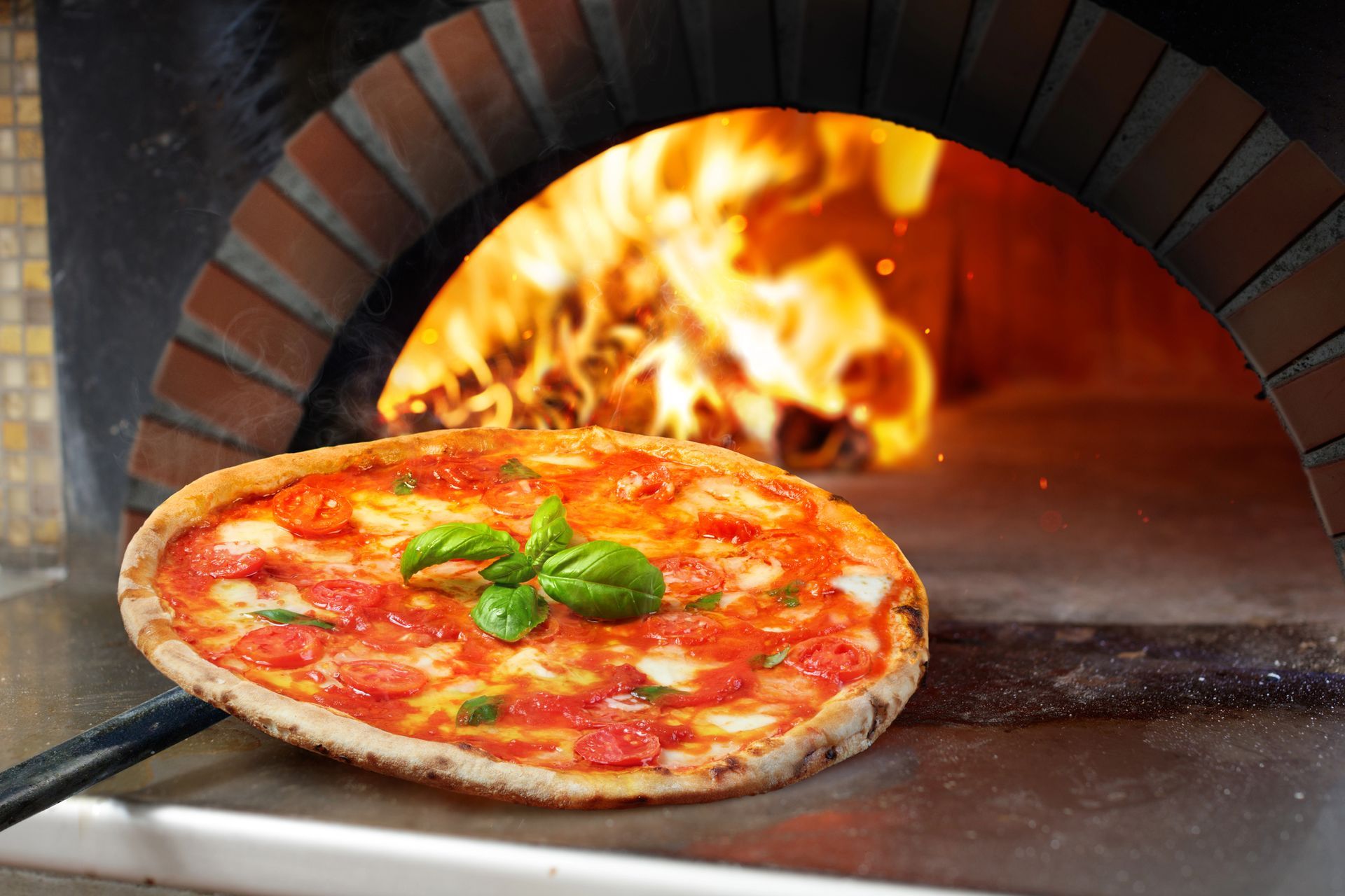 Una pizza Margherita appena sfornata con basilico fresco è adagiata su una pala di metallo davanti a un forno a legna in mattoni con le fiamme.