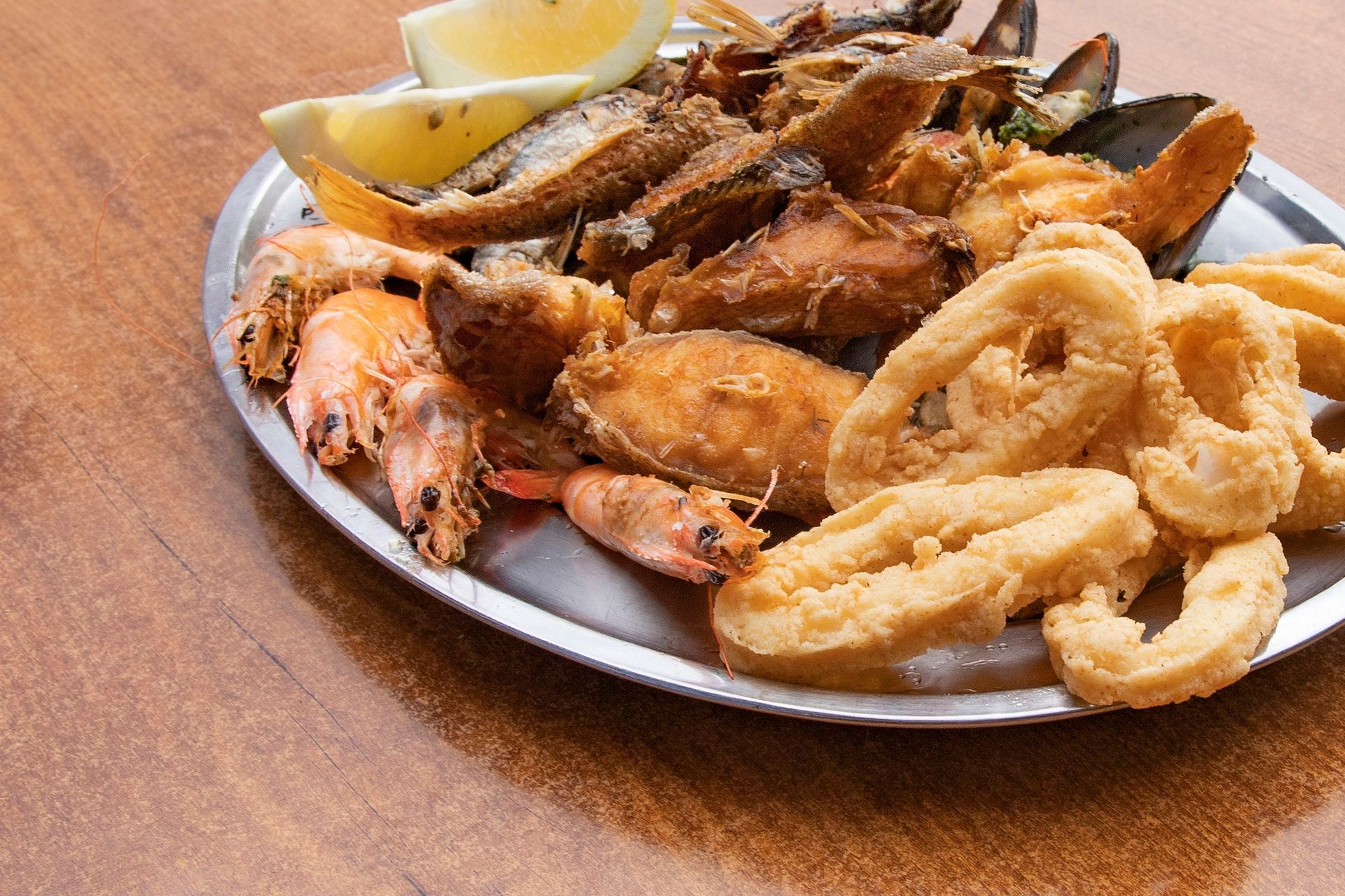 Un vassoio di metallo con una varietà di frutti di mare fritti, tra cui gamberi, calamari e cozze, guarnito con spicchi di limone.