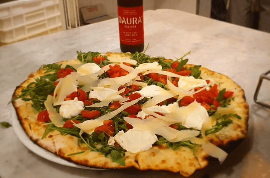 Una pizza condita con rucola, pomodori e scaglie di formaggio su un tavolo, con una bottiglia di birra sullo sfondo.