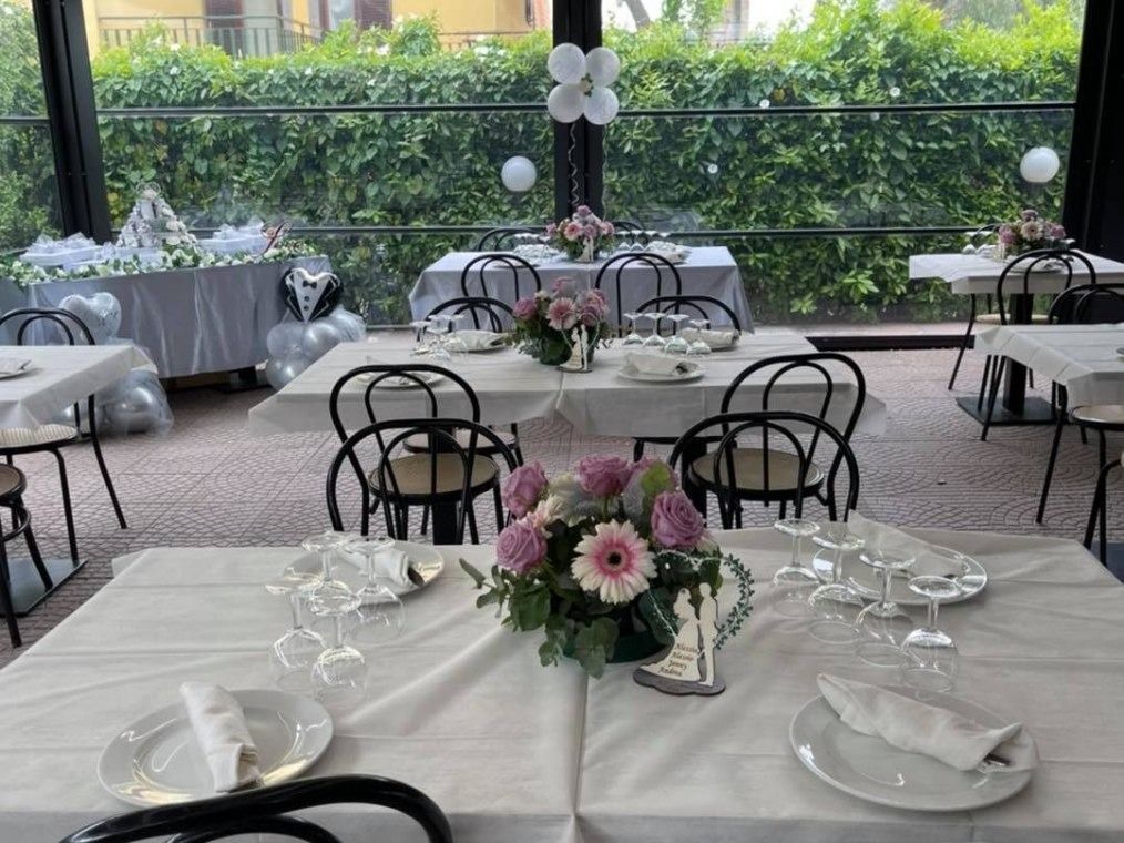 Tavoli apparecchiati per un evento formale in un padiglione interno illuminato dal sole, decorati con centrotavola floreali e palloncini bianchi.