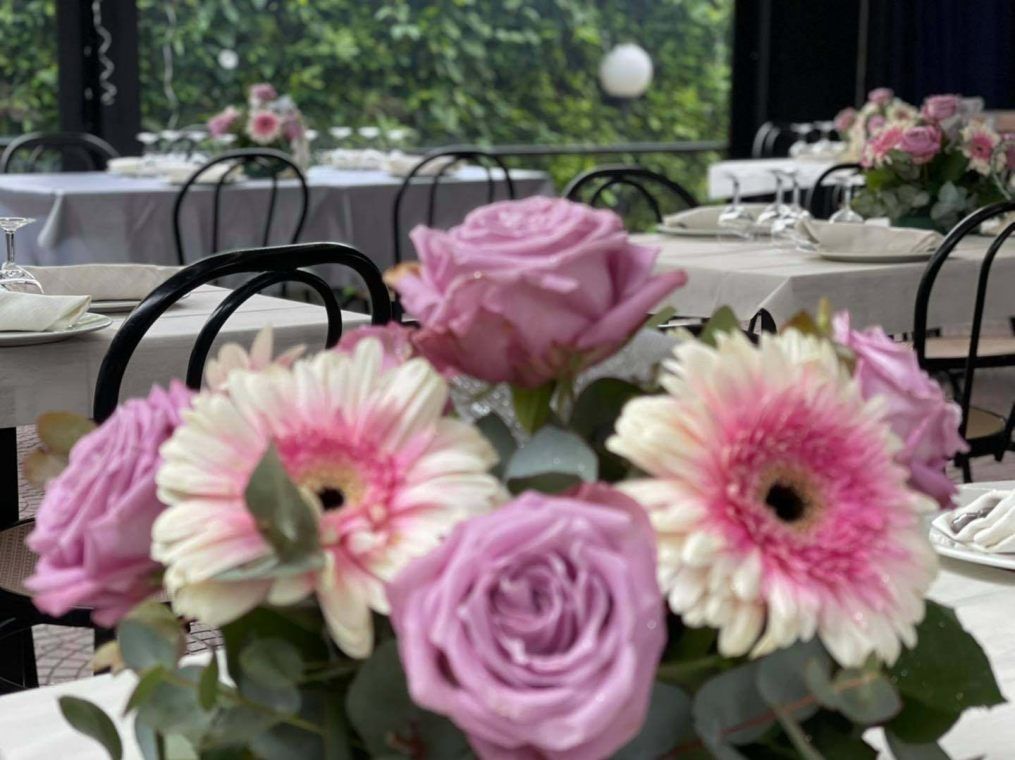 Un centrotavola composto da gerbere rosa e rose color lavanda, con tavoli e sedie sfocati sullo sfondo.
