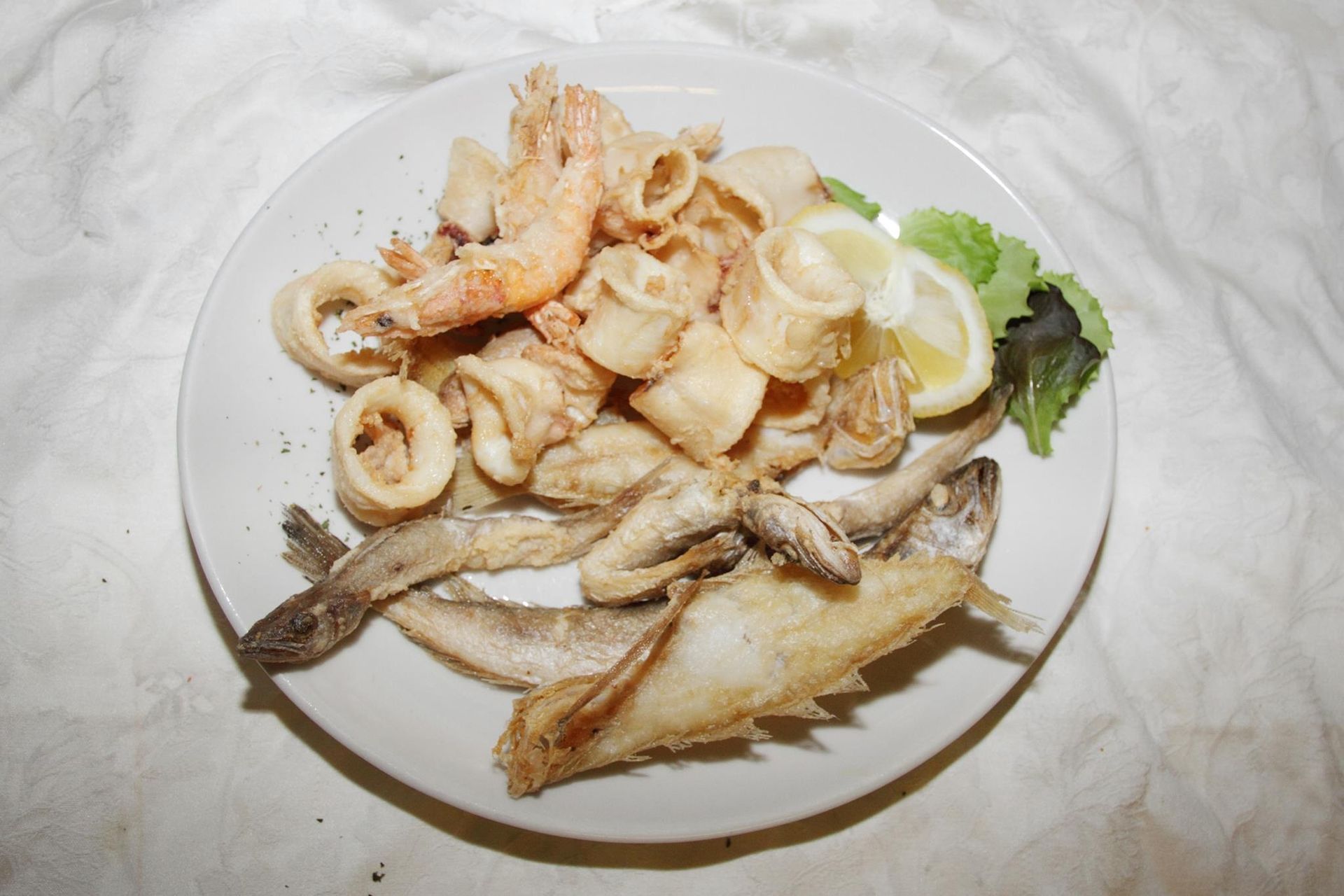 Un piatto di frutti di mare fritti, tra cui anelli di calamari dorati, gamberi e piccoli pesci interi, guarnito con limone e verdure.