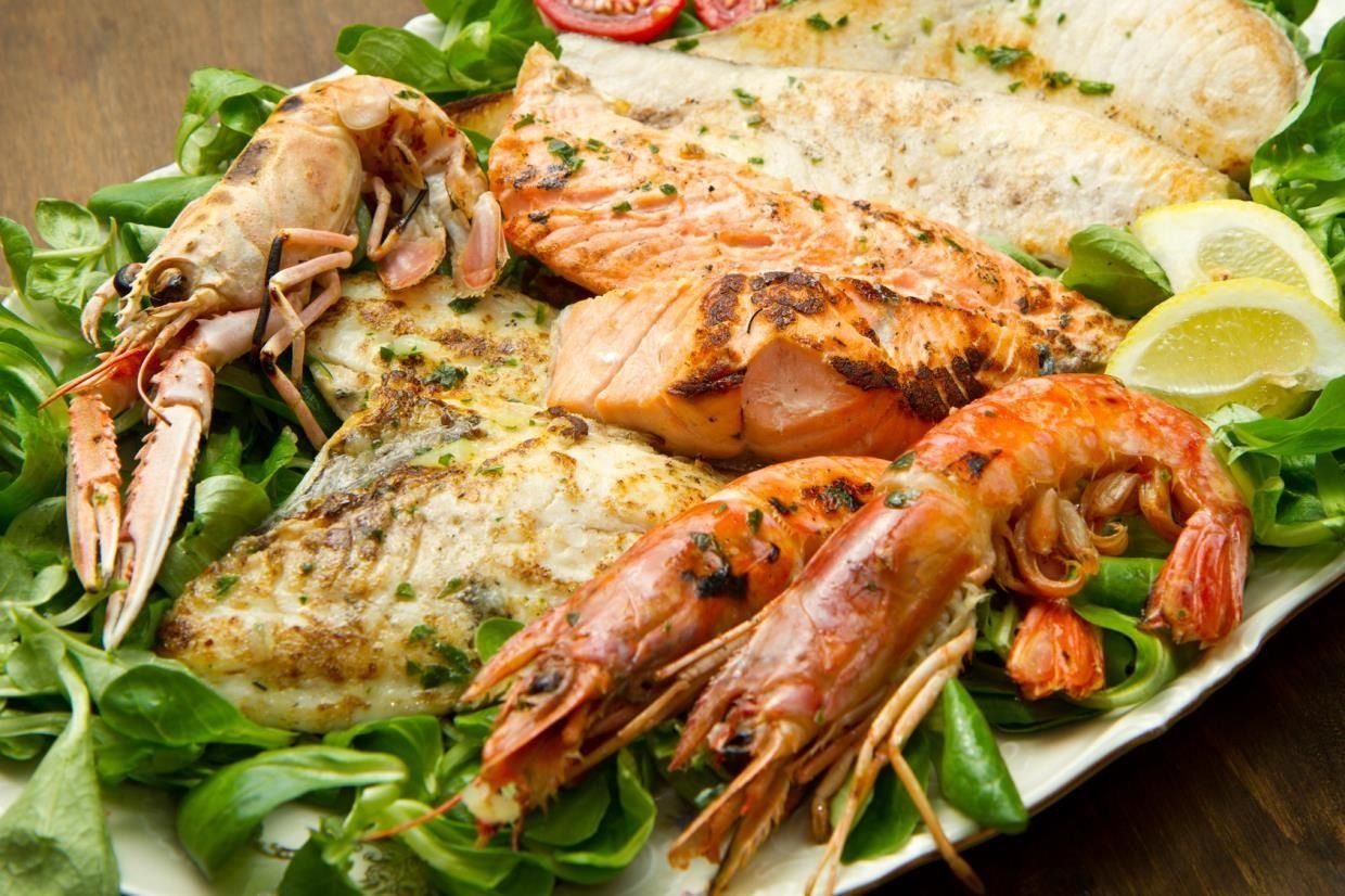 Un piatto misto di pesce grigliato, filetti di salmone e gamberoni, servito su un letto di verdure fresche con spicchi di limone.