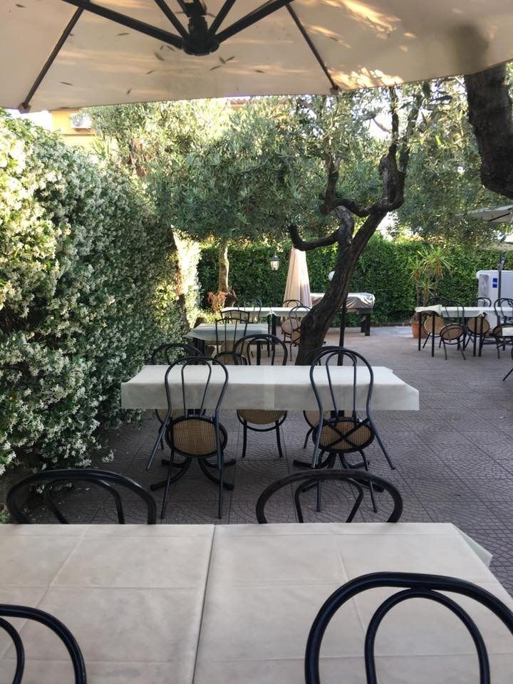 Dehor esterno del ristorante con tavoli, sedie e tovaglie bianche, riparato da un ombrellone tra alberi e vegetazione.