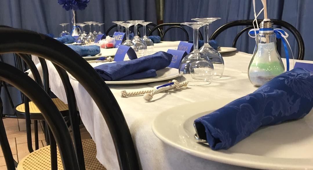 Una tavola apparecchiata per un evento formale con tovaglie bianche, tovaglioli blu scuro, bicchieri e centrotavola decorativi con lampadine.