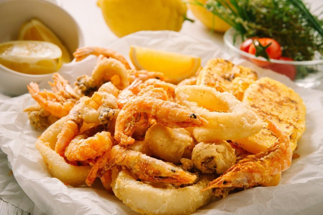 Un piatto di frutti di mare fritti, tra cui gamberi e calamari, dorati e croccanti, servito su carta da forno con spicchi di limone.