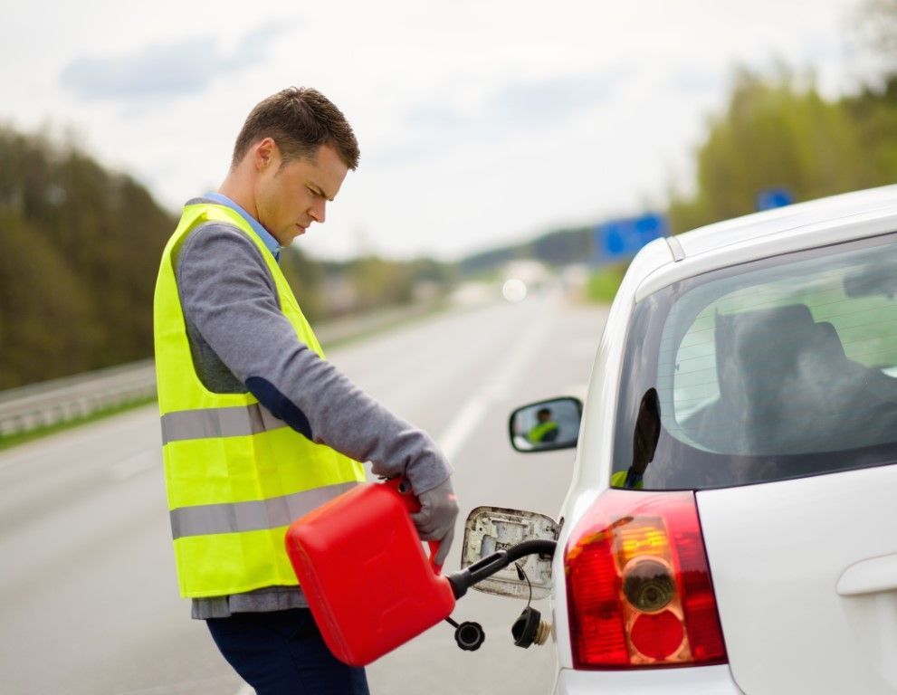 Man in a yellow vest pouring gasoline from a red can into a white car on the side of a highway.