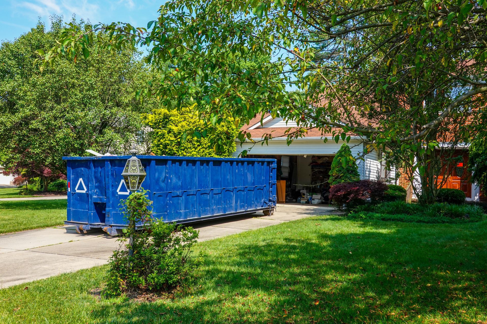 An empty blue dumpster in the driveway of a house with its garage door open.