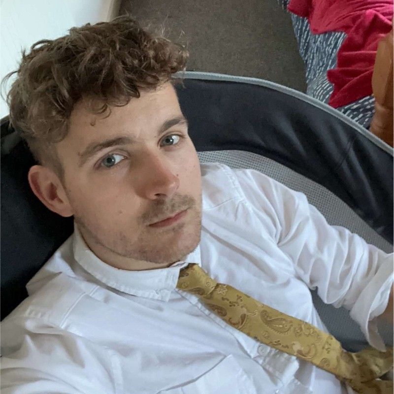 Image of Luke Rickards, with light eyes, and a white shirt, sitting in a chair with a gold tie, looking at the camera.