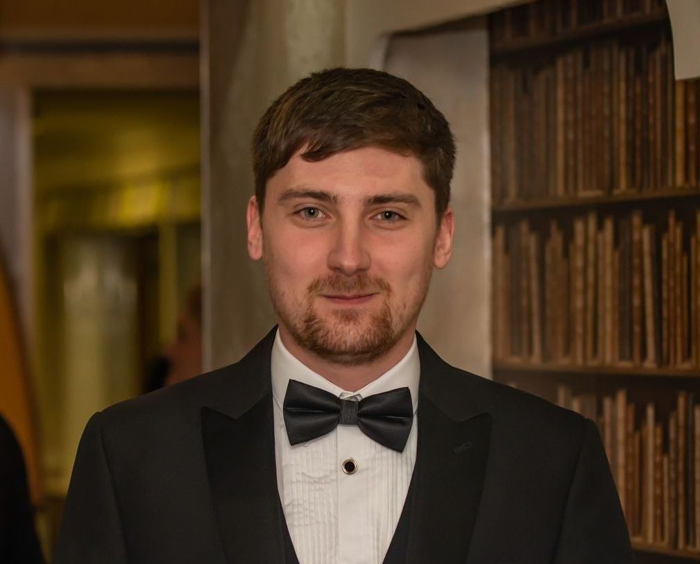 Image of Luke Statham in black tuxedo and bow tie, brown hair, slight beard, smiling, bookshelves in background.