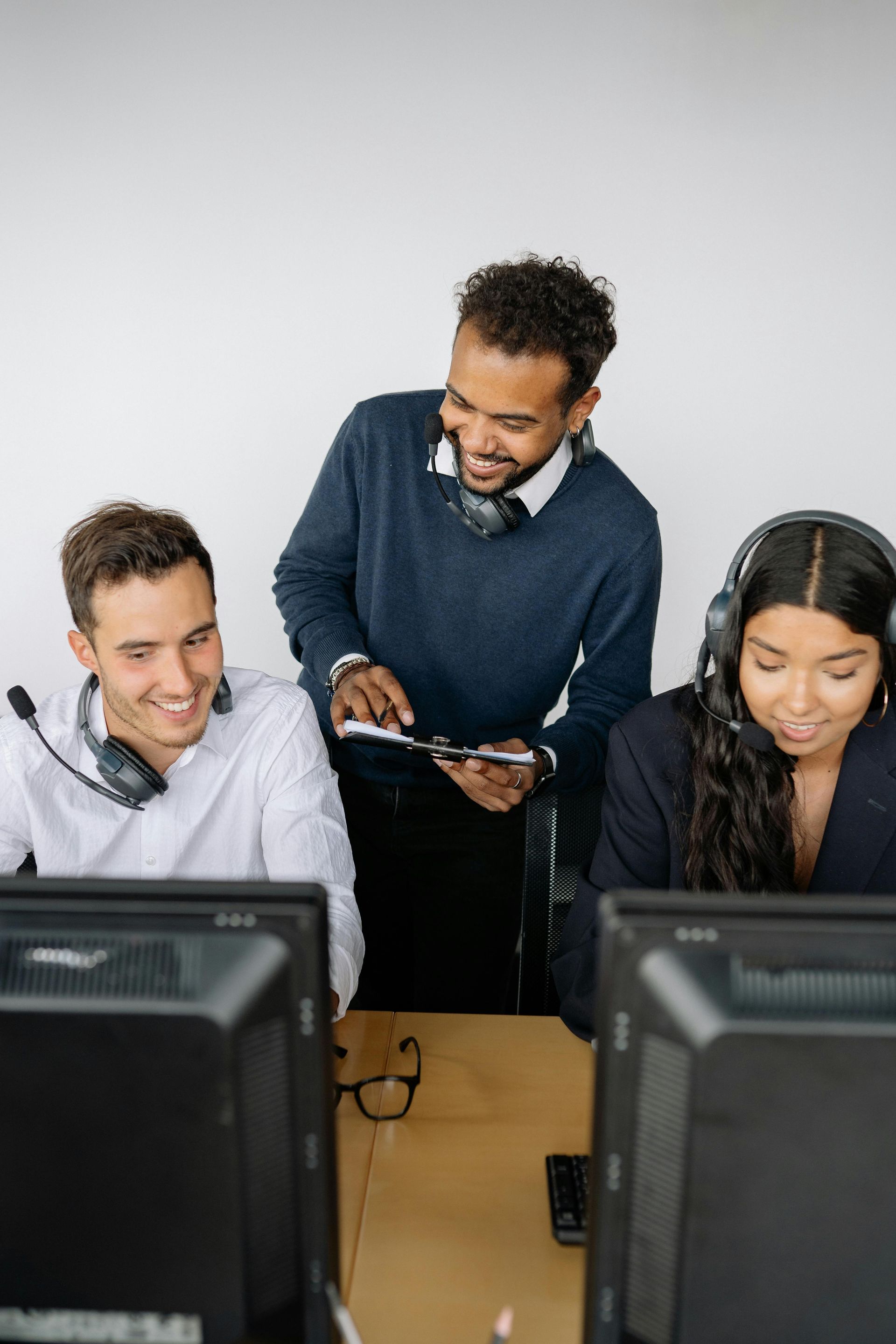 Three people at computers with headsets. One person stands, looking at a computer while smiling.