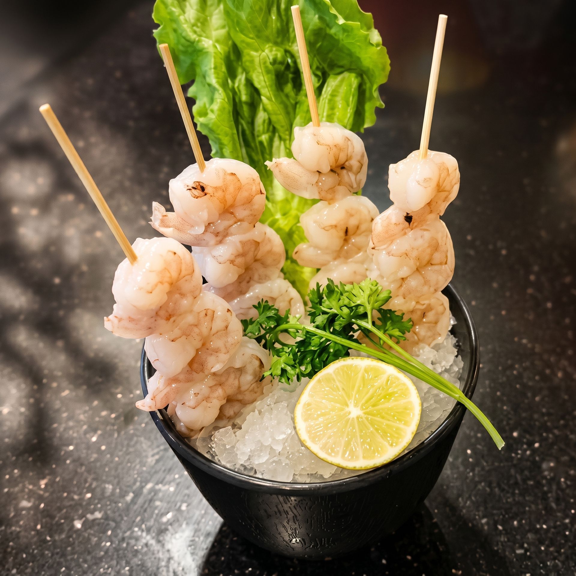 Shrimp skewers in a black bowl with ice, lemon slice, and greens on a dark table