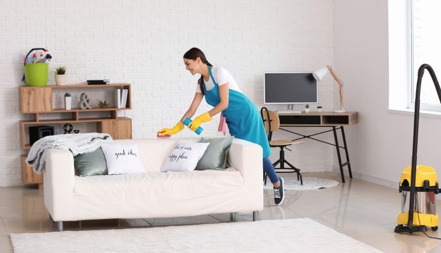Woman wearing gloves and apron cleans a white couch in a bright living room; vacuum cleaner nearby.