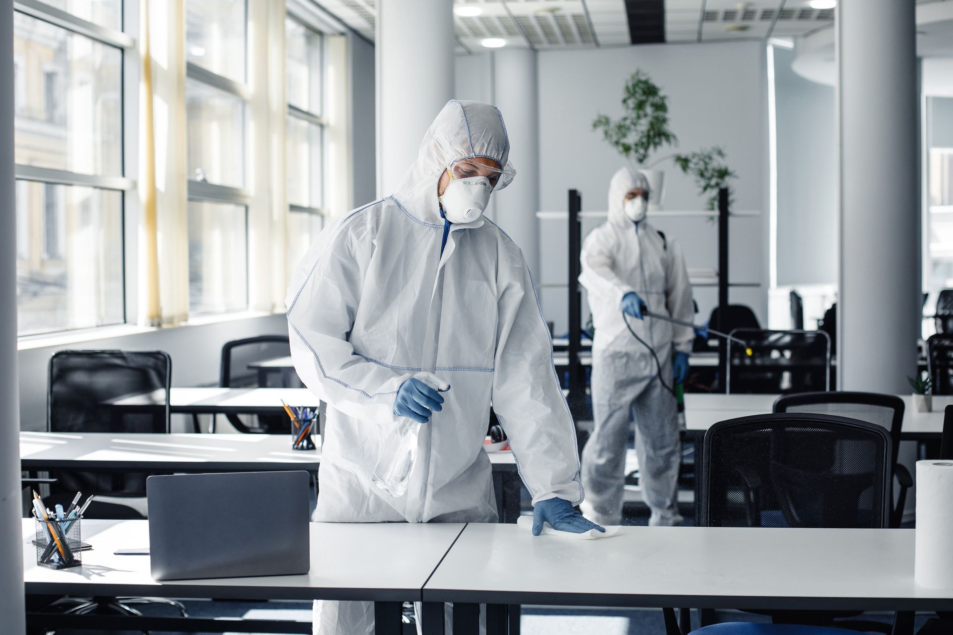 Two men in protective suits are disinfecting tables in an office.