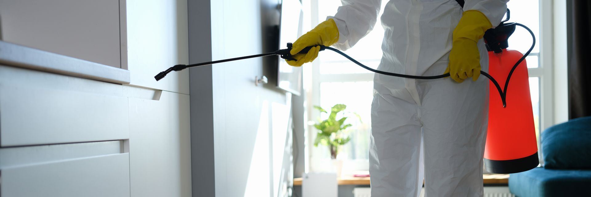 Person in protective suit spraying a white cabinet. Yellow gloves, red tank, interior.