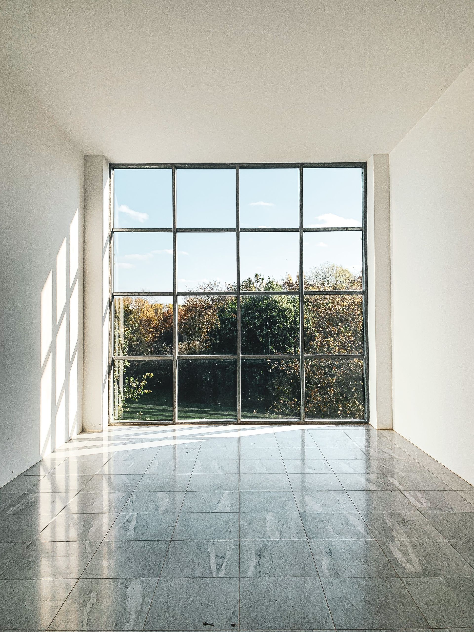 Empty room with a large, grid-style window overlooking a garden. Sunlight streams in, illuminating the polished floor.