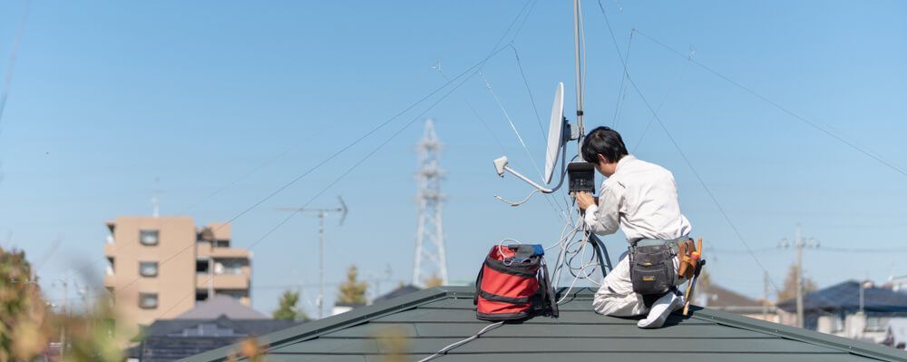 A Man Is Kneeling on Top of A Roof Working on A Satellite Dish — BTV Antenna & Data in Burnett Heads, QLD