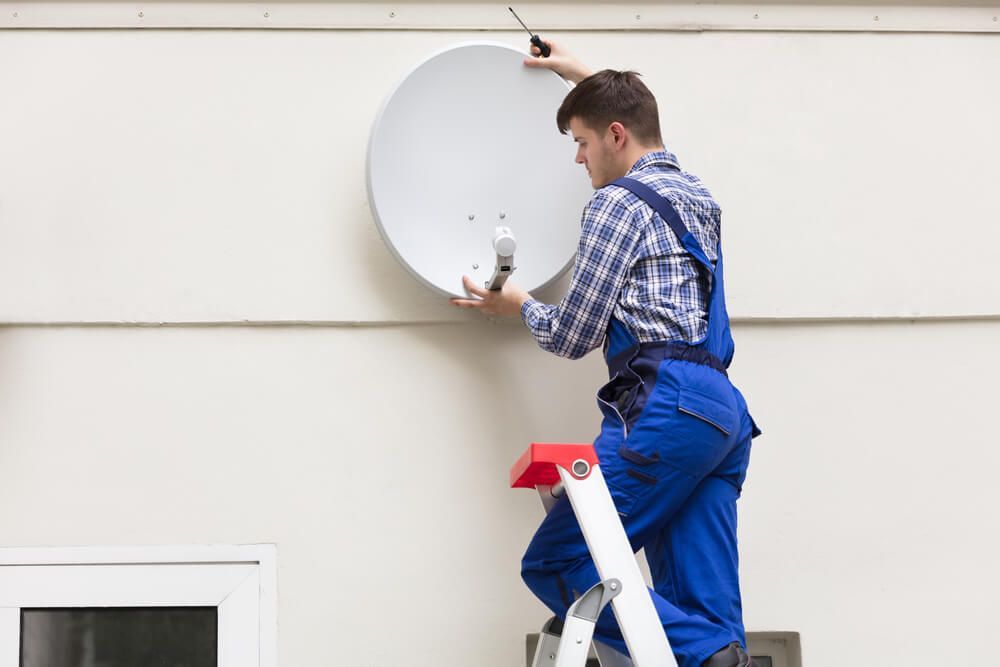 A Man Is Standing on A Ladder Fixing a Satellite Dish on A Wall — BTV Antenna & Data in Moore Park, QLD