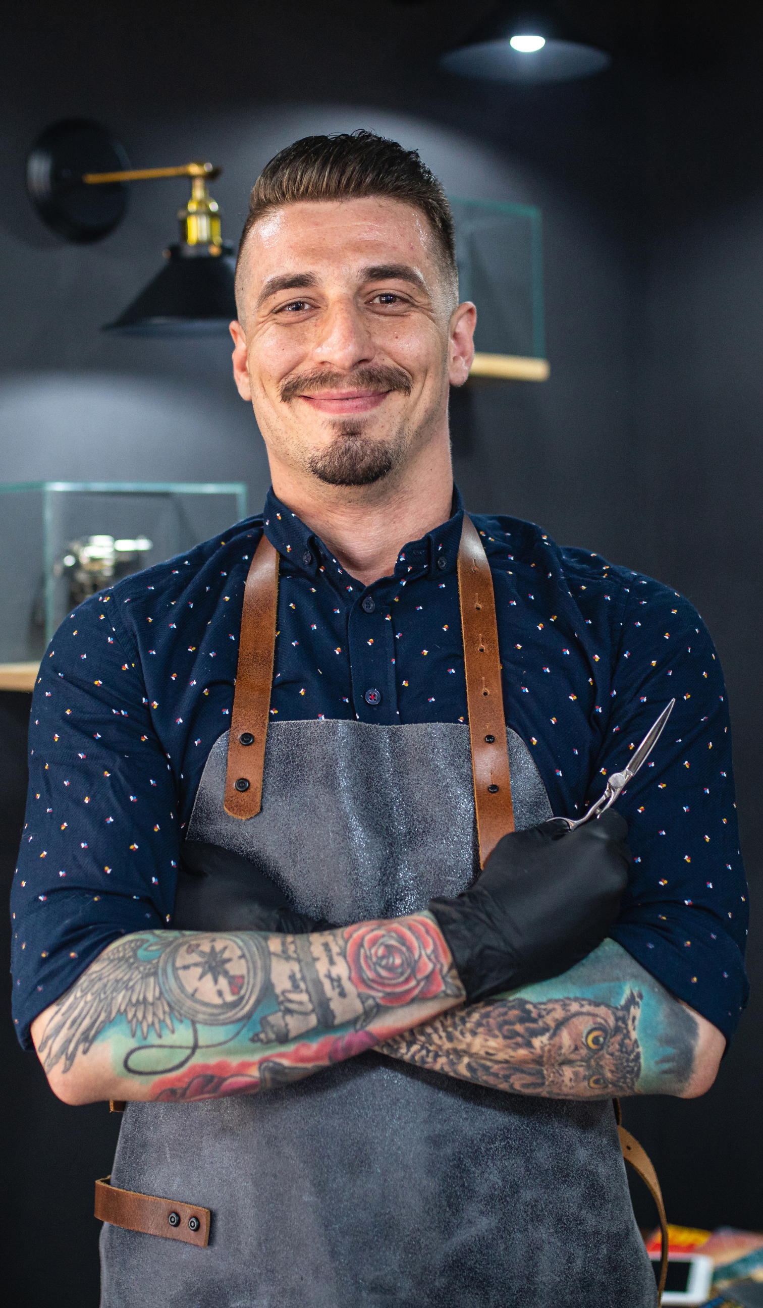 a barber wearing an apron and black gloves is standing with his arms crossed in a barber shop .