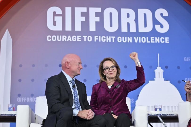 Gabby Gifford and her husband are sitting on stage at the annual conference  in front of a sign that says giffords courage to fight gun violence